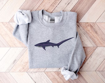 Könnte beinhalten: Ein graues Sweatshirt mit einem schwarzen und violetten Hai-Motiv auf der Vorderseite. Der Hai hat einen langen, dünnen Schwanz.