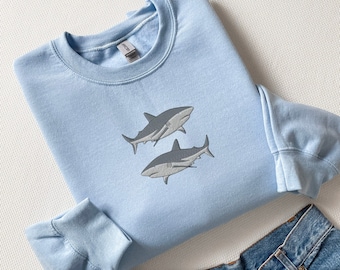 Pode incluir: Uma camisola de gola redonda azul-clara com um design bordado de dois tubarões cinzentos a nadar no centro. A camisola tem mangas compridas com punhos canelados e uma gola canelada. Umas calças de ganga azuis são visíveis na parte inferior.