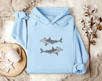 Puede incluir: Una sudadera con capucha de color azul claro que presenta un diseño bordado de dos tiburones martillo grises colocados uno encima del otro en el centro del pecho. La prenda incluye una capucha con cordón y un bolsillo canguro frontal.