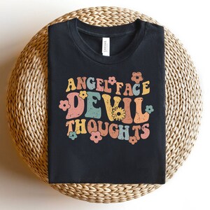 Könnte beinhalten: Schwarzes T-Shirt mit einer Grafik im Retro-Stil, die "Angel Face Devil Thoughts" in bunten Buchstaben mit Blumen zeigt.