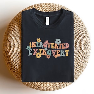 Może przedstawiać: Czarny T-shirt z grafiką w stylu retro, na której widnieje napis "Introvert Ekstrawertyk" otoczony kolorowymi kwiatami.