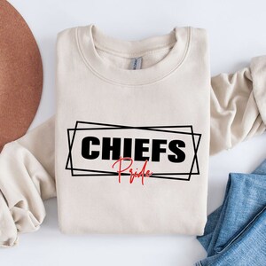 May include: A beige sweatshirt with the words "CHIEFS Pride" printed in black and red lettering inside a black outlined box.