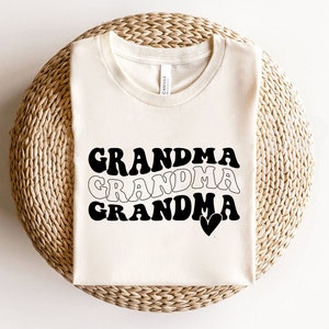 May include: A white t-shirt with the text "Grandma" written three times in black, with each word in a slightly different retro font. The last word has a small heart next to it.