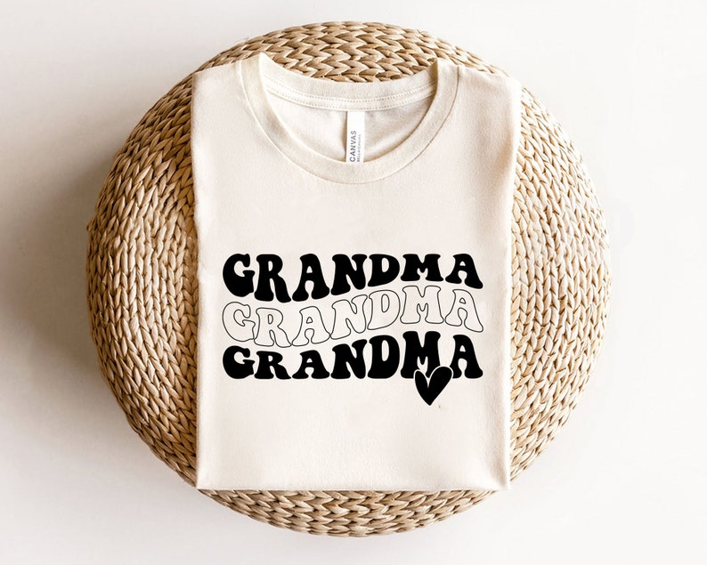 May include: A white t-shirt with the text "Grandma" written three times in black, with each word in a slightly different retro font. The last word has a small heart next to it.