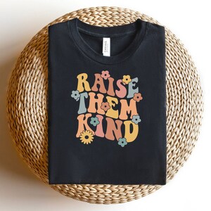 Könnte beinhalten: Schwarzes T-Shirt mit einer Retro-Grafik, die "Raise Them Kind" in bunten Buchstaben mit Blumen zeigt.