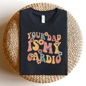 May include: Black t-shirt with a retro-style graphic that says "Your Dad Is My Cardio" with colorful flowers surrounding the text.