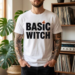 Può includere: Una t-shirt bianca a maniche corte con la scritta nera BASIC WITCH stampata sul davanti. Una piccola icona di un cappello da strega arancione è posizionata sopra la lettera I nella parola BASIC. La maglietta ha un girocollo e una vestibilità standard.