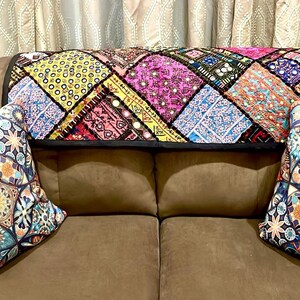 May include: A brown couch with two pillows featuring a colorful geometric pattern. A colorful patchwork throw with sequins and mirrors is draped across the back of the couch.