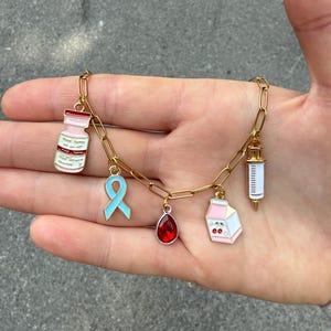 Op de afbeelding: Een goudkleurige kettingarmband met meerdere emaille bedels. Bedels zijn onder meer een roze en witte medicijnfles, een lichtblauw lint, een rode druppel, een roze melkpak met kersen en een injectiespuit.