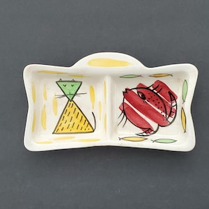Puede incluir: Un plato de cerámica dividido con un diseño caprichoso. Un lado presenta un gato amarillo con cabeza verde, el otro un pez rojo. El plato es blanco con detalles amarillos, verdes y negros, ideal para servir aperitivos o porciones pequeñas.