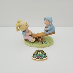 Può includere: Una statuetta in porcellana di due Cabbage Patch Kids su un'altalena. La ragazza indossa un vestito rosa e il ragazzo indossa un vestito blu. La statuetta è su una base verde con fiori e il logo Cabbage Patch Kids.