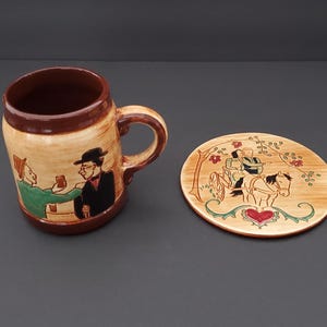 May include: A ceramic mug and coaster set. The mug features a brown rim and handle, with a tan body depicting two men toasting. The coaster shows a horse-drawn carriage with a heart design. A charming, vintage-style set.