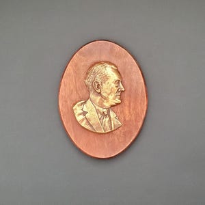 May include: An oval-shaped wooden plaque featuring a raised, metallic gold-toned relief sculpture of a person in profile. The figure is depicted from the chest up, wearing a collared shirt, tie, and suit jacket, set against a smooth, reddish-brown wood.