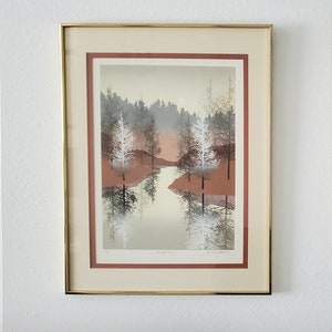 Könnte beinhalten: Gerahmter Druck einer Landschaftsszene mit einem Fluss, der durch einen Wald fließt. Die Bäume sind in einem gedämpften Grün und Braun gehalten, und das Wasser ist hellblau. Der Druck ist in einem goldenen Rahmen mit einer roten Passepartout.