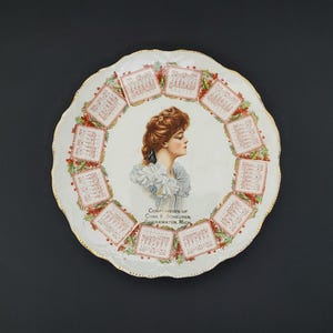 May include: A white ceramic decorative plate with a scalloped gold-trimmed edge. The center features a portrait of a person with reddish-brown hair. Surrounding the portrait are twelve monthly calendar grids for 1909, accented with holly leaves.