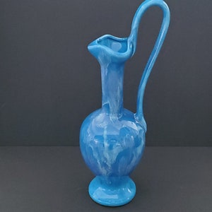 May include: A blue ceramic pitcher with a white marbled glaze and a curved handle. The pitcher has a round body and a flared base.