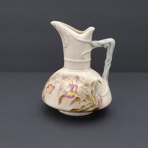 May include: A cream-colored ceramic pitcher featuring a bulbous base decorated with painted orchid flowers in shades of pink, yellow, and green. The vessel has a curved spout and a textured, twisted handle that extends from the neck to the body.