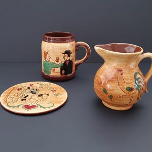 Könnte beinhalten: Ein Keramikset bestehend aus einem braun-beigen Bierkrug mit einer bemalten Szene von zwei Personen, einem runden Teller mit einer Pferde- und Reiterillustration sowie einem kleinen Krug mit Hahn-Motiv in Braun-, Grün- und Rottönen.