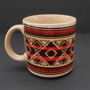 May include: A ceramic coffee mug with a cream-coloured handle and interior. The exterior features a geometric pattern in red, black, and tan. The design includes diamond and zigzag shapes. The mug is suitable for hot or cold beverages.