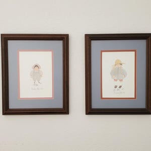 May include: Two framed illustrations of children in vintage clothing. The illustrations are in a watercolor style and feature a girl wearing a straw hat and a boy wearing a jacket. The frames are brown wood with a light blue mat.