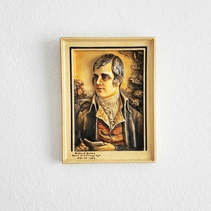 May include: A framed portrait of Robert Burns, the Scottish poet, in a cream-colored frame. The portrait is in a relief style, with a textured surface. The text below the portrait reads "Robert Burns, born at Alloway Ayr, Jan. 25, 1759."