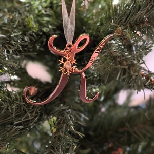 May include: A rose gold letter K Christmas bauble with a snowflake charm. The bauble is hanging on a green artificial Christmas tree.