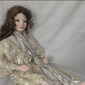 May include: A porcelain doll with brown hair and blue eyes, wearing a gold and silver patterned gown and a matching scarf. The doll is posed in a seated position, with delicate facial features and a serene expression. The doll's attire has a shimmering, elegant appearance.