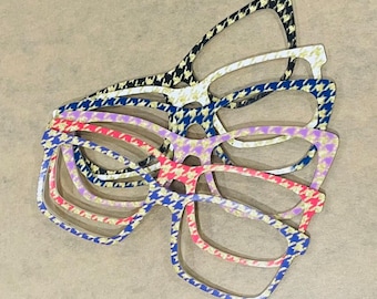 May include: A stack of eyeglasses frames in various colors, including black and white, blue and white, pink and white, and purple and white. Each frame features a houndstooth pattern.
