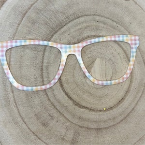 May include: A pair of oversized glasses with a pastel gingham pattern. The glasses are made of a white material with a light pink, yellow, and blue checkered design.