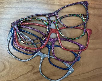 May include: A collection of glitter-covered eyeglasses in various colors, including purple, red, blue, and green. The frames are stacked on a wooden surface, showcasing the different color options available.