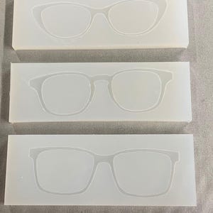 May include: Three white silicone molds, each with a different eyeglass frame design. The top mold features a cat-eye style, the middle a classic square shape, and the bottom a rectangular frame. These molds are likely for crafting or resin projects.