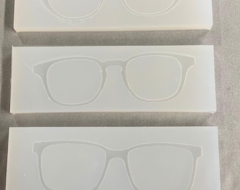May include: Three white silicone molds, each with a different eyeglass frame design. The top mold features a cat-eye style, the middle a classic square shape, and the bottom a rectangular frame. These molds are likely for crafting or resin projects.