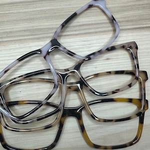 Può includere: Quattro paia di montature per occhiali impilate, che mostrano una varietà di colori e motivi. Le montature presentano un mix di bianco, nero e marrone, con un motivo tartarugato. Le montature sono realizzate in materiale lucido.