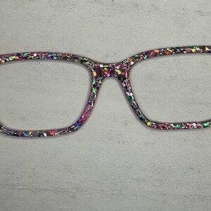 May include: A pair of square-shaped glasses with a glittery, iridescent frame. The frame is covered in small, colourful glitter.