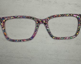 May include: A pair of square-shaped glasses with a glittery, iridescent frame. The frame is covered in small, colorful glitter.