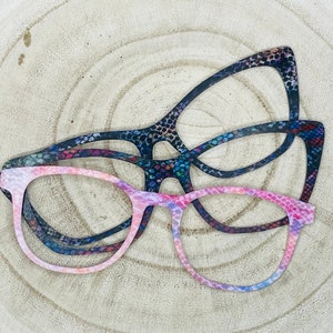 May include: Two pairs of spectacle frames with a snakeskin pattern. One pair is pink and purple, the other is dark blue and black. The frames are layered on a light-coloured, textured surface.