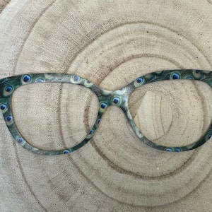 May include: A pair of black cat-eye glasses with a peacock feather print. The glasses have a blue and green pattern.