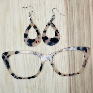 May include: A pair of tortoise shell earrings with a teardrop shape and a cutout design.  The earrings are hanging from silver hooks.  A pair of tortoise shell glasses with a cat eye shape.