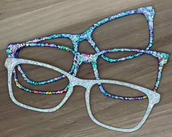May include: Two pairs of decorative eyeglasses with glitter. One pair is white with silver glitter, and the other is blue with a mix of colorful glitter. The glasses are rectangular shaped and appear to be made of a lightweight material.