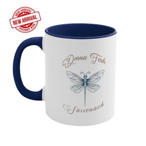 May include: A white ceramic mug with a dark blue interior and handle. The front features a detailed blue dragonfly illustration centred between the brown cursive text Dinna Fash above and Sassenach below. A red circular New Arrival badge is visible.