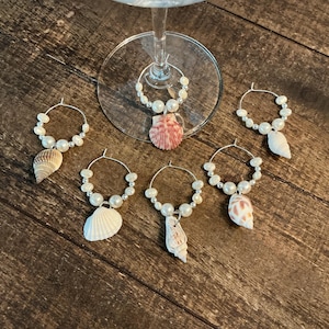 May include: A collection of six wine glass charms. Each charm features a silver hoop adorned with faux pearls and a seashell. The seashells vary in shape and color, including white, pink, and brown.