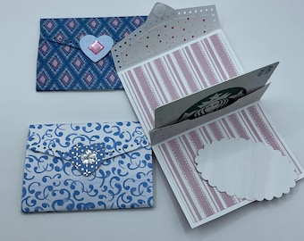 May include: Three handmade gift card holders. One is blue with a pink diamond pattern and a heart-shaped gem. Another is blue with a white swirl pattern and a heart-shaped gem. The third is open, showing a Starbucks gift card.