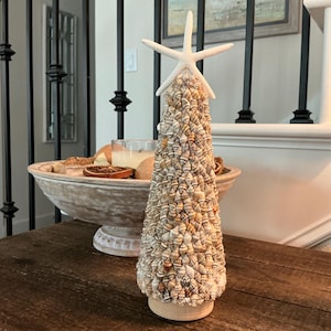 May include: A decorative seashell tree with a white starfish topper. The tree is made of various seashells in shades of tan, cream, and brown. It sits on a light-colored base. The tree is displayed on a dark wooden table.