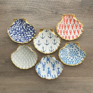 May include: A collection of six seashell-shaped trinket dishes. Each dish features a gold-painted rim and a unique interior design, including anchors, lobsters, and floral patterns in blue, white, and red.