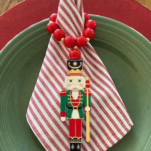 May include: A festive table setting featuring a red and white striped napkin held by a red beaded napkin ring. A decorative nutcracker figurine stands on the napkin, placed on a green plate atop a red charger plate. Holiday themed.