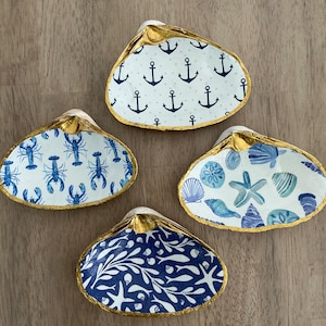 May include: Four decorative seashell dishes with gold-painted edges. Each shell features a different nautical design: anchors, lobsters, seashells, and a blue and white pattern. The shells are arranged on a wooden surface.