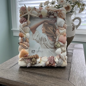 May include: A decorative photo frame adorned with a variety of seashells, including a starfish, in shades of white, beige, and pink. The frame holds a 5x7 inch (12.7cm x 17.7cm) photo. The frame is placed on a wooden surface.