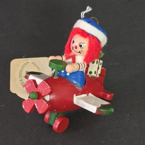 May include: A red wooden airplane ornament with a white propeller and green wheels. A red-haired doll with a blue and white sailor hat is sitting in the cockpit holding a green steering wheel. The doll is carrying a green and white gift box.