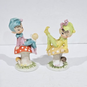 May include: Two ceramic figurines of elves sitting on mushroom-shaped bases. The elf on the left is wearing a blue and pink outfit and has a snail on its shoulder. The elf on the right is wearing a yellow and green outfit and has a pink worm on its head.