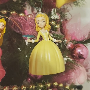 May include: A yellow princess ornament with a crown, holding a green leaf. The ornament is on a Christmas tree with pink and gold decorations.